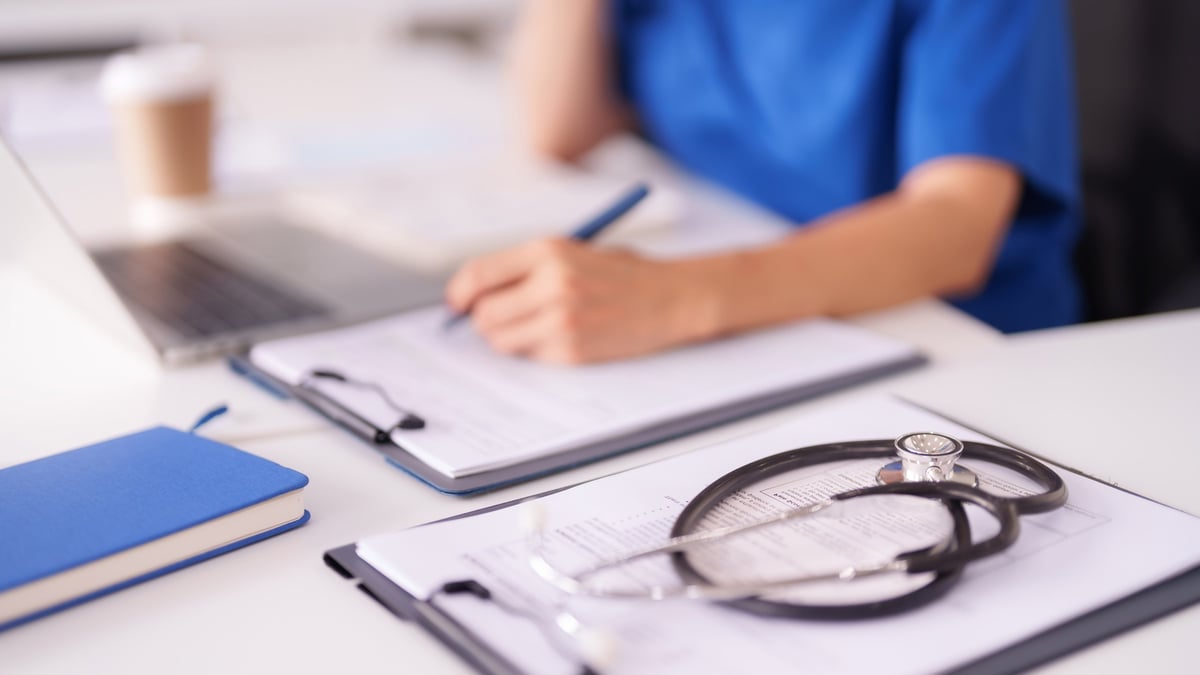 Adult healthcare worker records notes on clipboard with laptop stethoscope clinic office desk professional medical environment